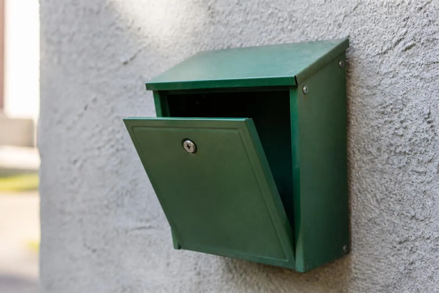 Une boîte aux lettres métallique verte entrouverte et vide, accrochée à un mur de crépi gris.